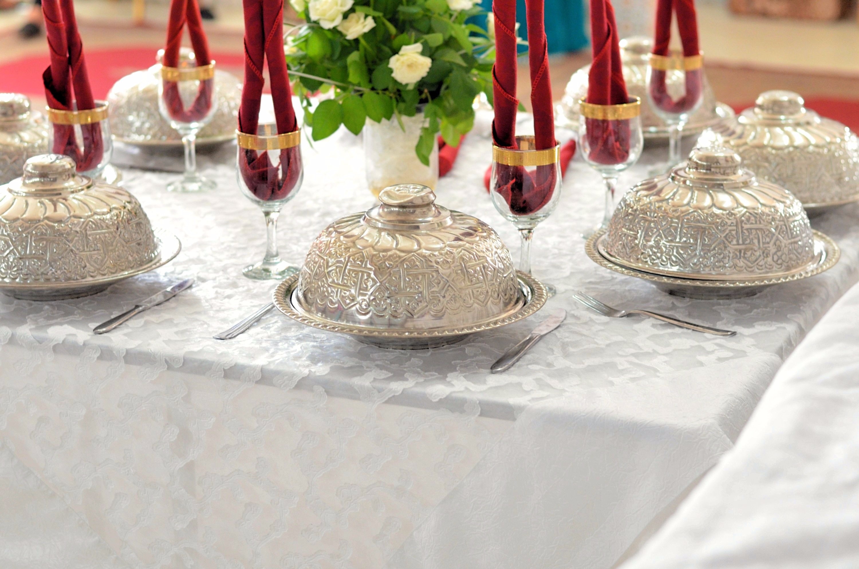 Ornate silver plate covers and stemmed glasses with folded cloth napkins are set on a table covered by white linen.