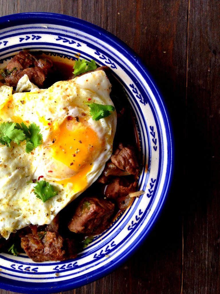 Kebab maghdour topped with fried eggs and served in a white tagine with blue patterns