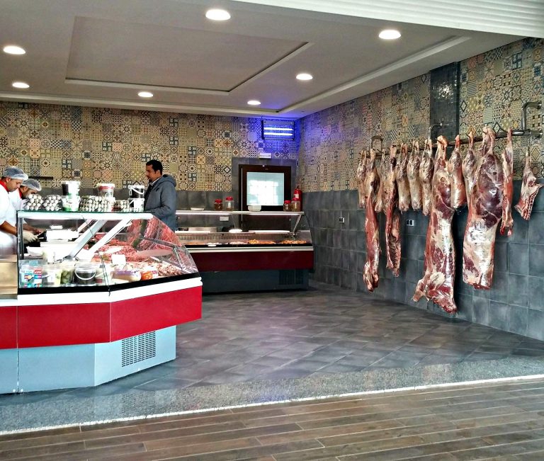 image of a Moroccan bucher shop. A customer is standing next to a glass-enclosed meat counter on the left and fresh whole meat is hanging to age on the right.