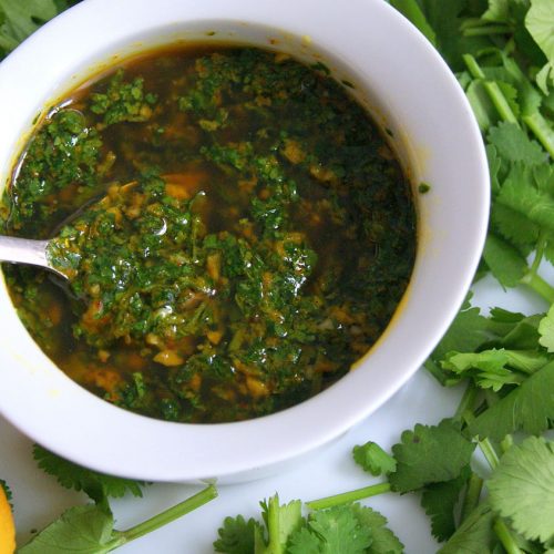 Chermoula marinade in a white bowl, topped with olive oil and surrounded by coriander leaves. A lemon appears in the corner.