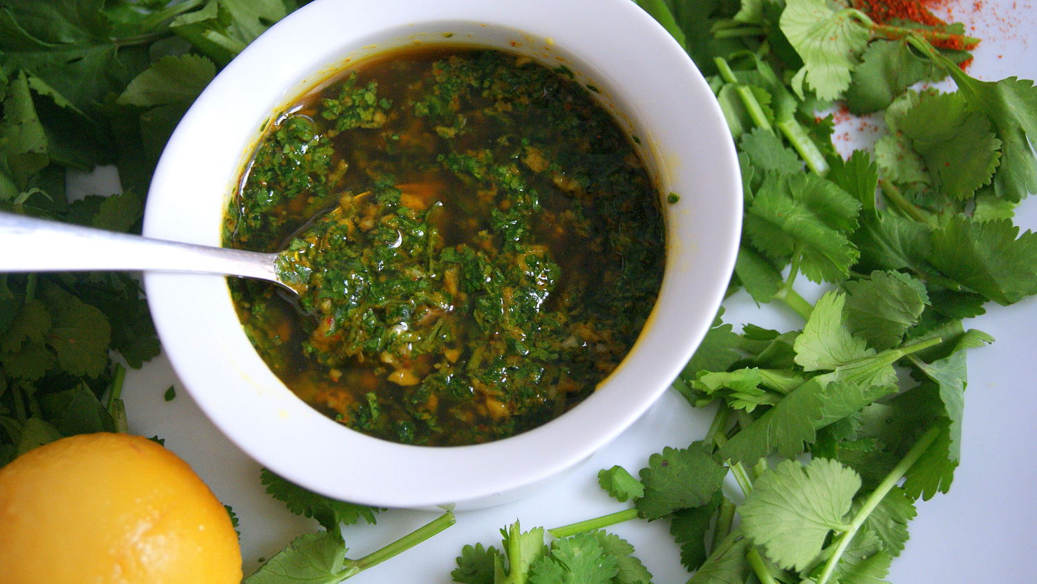Chermoula marinade in a white bowl, topped with olive oil and surrounded by coriander leaves. A lemon appears in the corner.