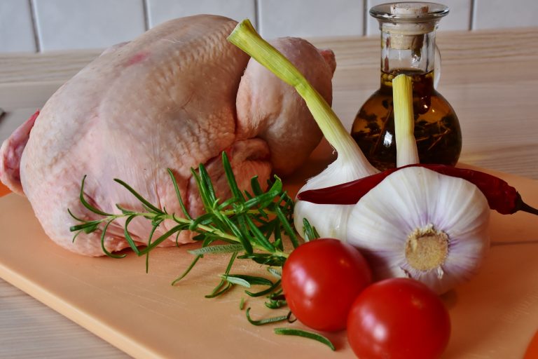 Fresh whole chicken on a wooden cutting board. Two heads of garlic, two tomatoes and a sprig of rosemary are beside the chicken.