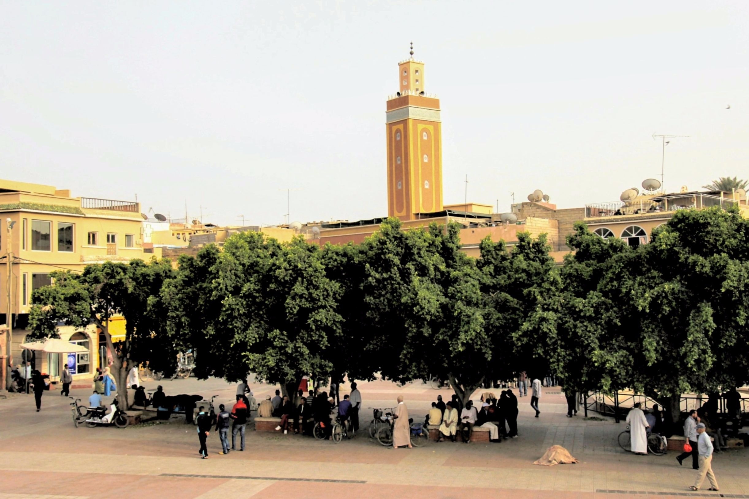 Image of garden square in Essaouira, Morocco. A mosque is in the background and people are walking and milling about, but the perspective is too far away to make out faces.