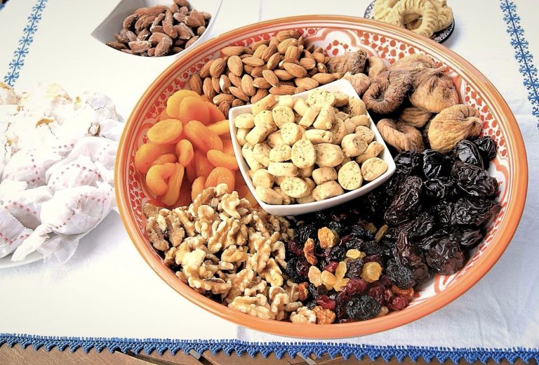A large decorated serving platter features a bowl of krichlate, miniature shortbread cookies surrounded by almonds, walnuts, dried figs, dried apricots, dates and raisins.
