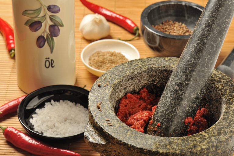 Close-up view of a mortar and pestle alongside chili peppers, coarse salt and spices. Harissa chili paste can be seen in the mortar.