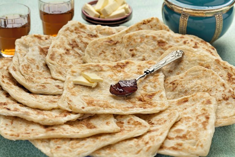 Square, laminated pancakes called msemen are arranged in overlapping fashion on a platter. Two pats of butter and a small spoon full of jam sits on the pile of msemen.