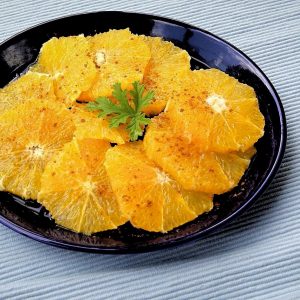 Round orange slices are arranged in overlapping fashion on a deep blue plate. The oranges are garnished with ground cinnamon.