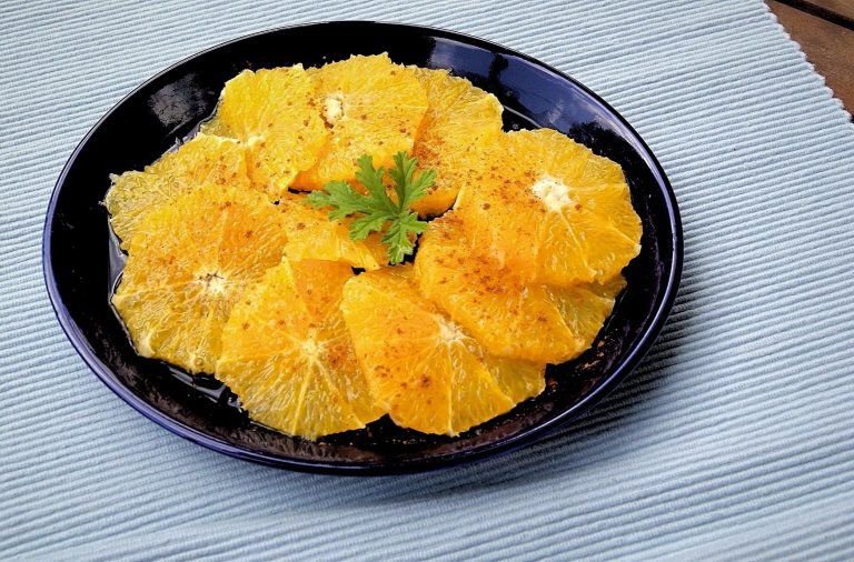 Round orange slices are arranged in overlapping fashion on a deep blue plate. The oranges are garnished with ground cinnamon.