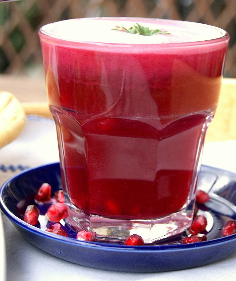 Moroccan pomegranate juice in a glass. The glass is on a small blue saucer and is surrounded by pomegranate seeds