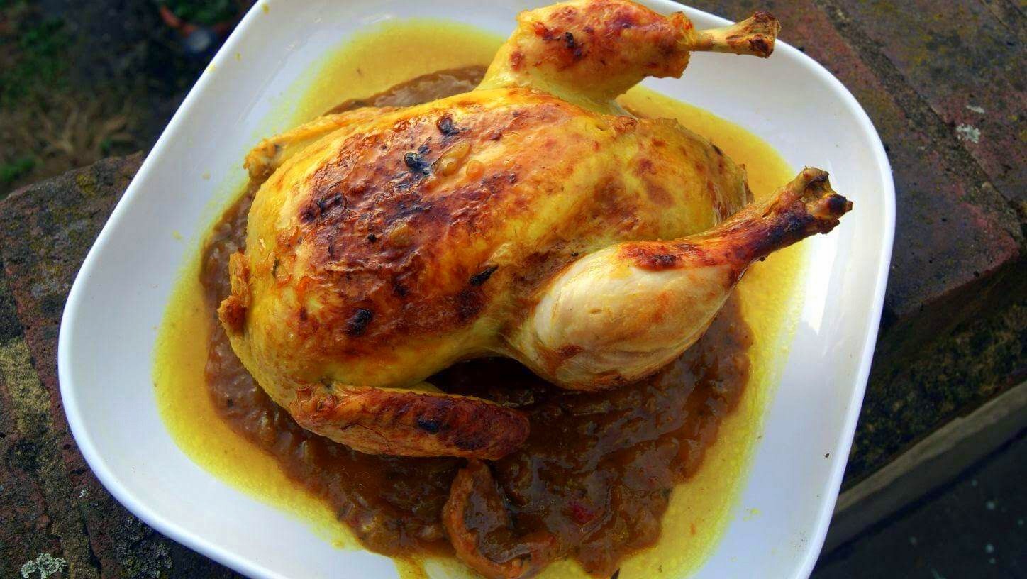 A whole chicken prepared m'qualli style sits on a plate surrounded by rich onion sauce which has separated from the oil. The chicken's skin is visibly roasted from a final stage of cooking in the oven.