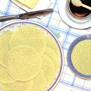 Overhead view of Moroccan semolina pancakes with a honeycombed appearance. The beghrir are arranged in overlapping fashion on the serving plate. A small plate holds a single pancake. Butter and honey can bee seen on the edge of the photo.