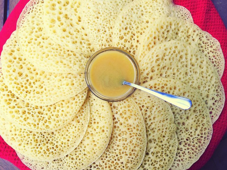 Overhead view of spongy semolina pancakes called beghrir. The pancakes have a honeycombed appearance and are arranged in an overlapping circular pattern on the serving dish. A small bowl of butter and honey syrup is centered on the pile of pancakes.