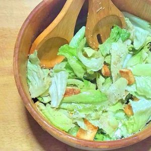 Overhead view of a wooden salad bowl filled with Caesar salad: crisp green romain lettuce topped with homemade croutons and shaved Parmesan cheese. Large wooden salad spoon and fork are resting in the bowl.
