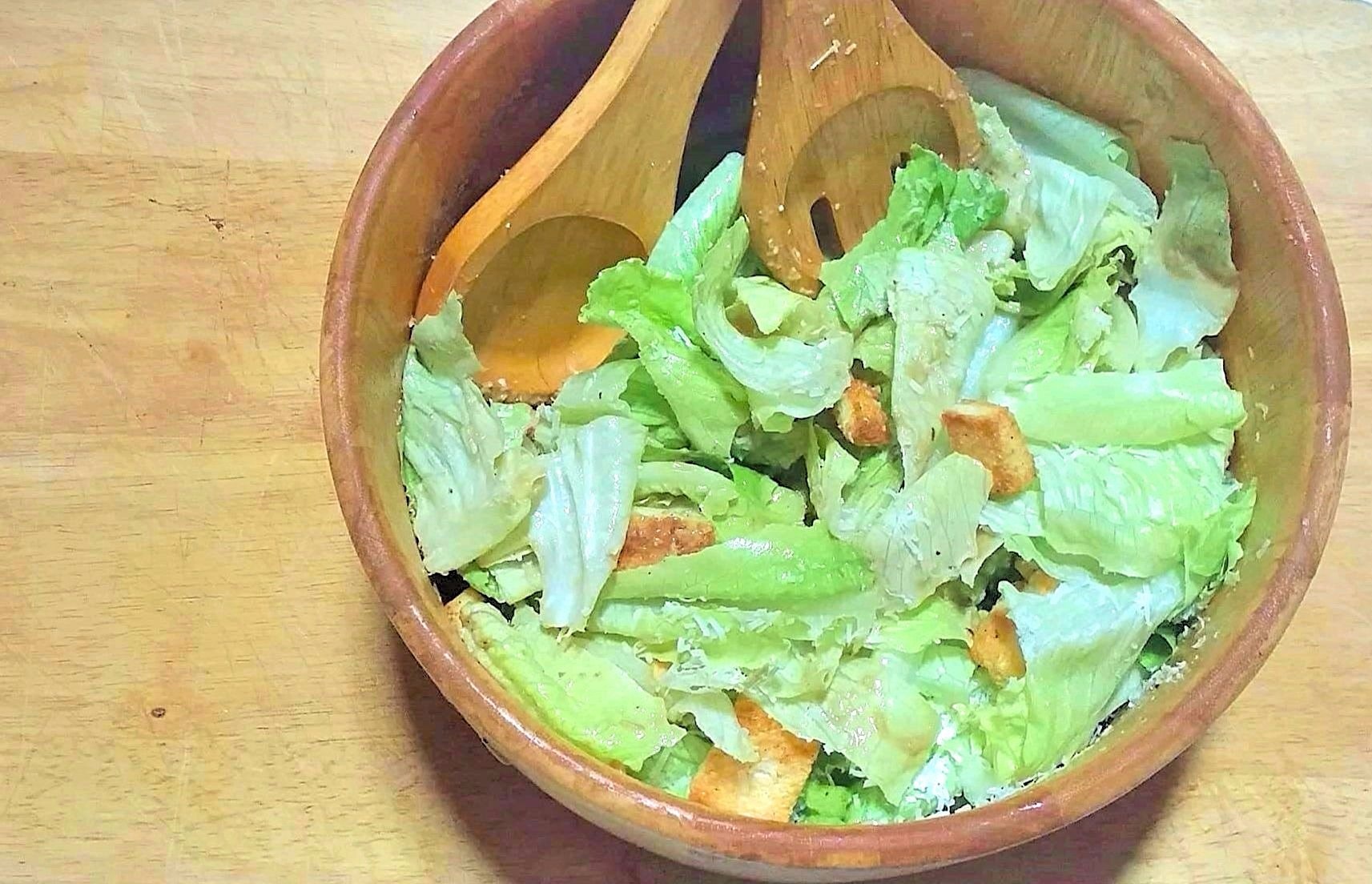 Overhead view of a wooden salad bowl filled with Caesar salad: crisp green romain lettuce topped with homemade croutons and shaved Parmesan cheese. Large wooden salad spoon and fork are resting in the bowl.
