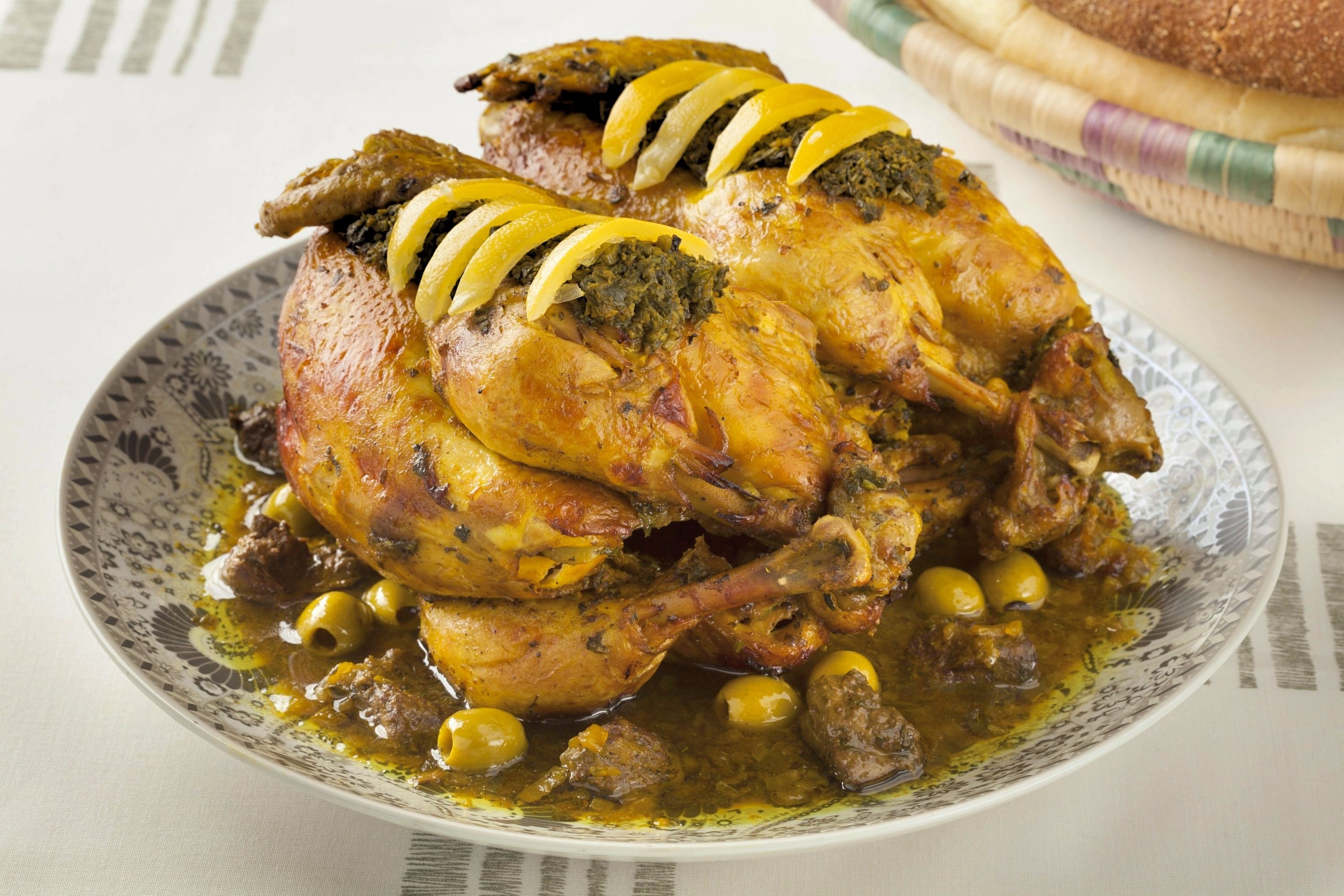 Two roasted chickens sit side-by-side on a serving platter. They are trussed and garnished with chermoula marinade and strips of preserved lemon skin. The chickens are surrounded by an onion sauce with chicken liver and green olives.