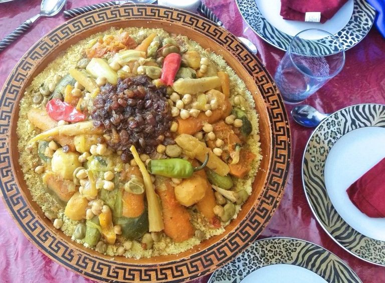 Overhead view of gorgeous presentation of Moroccan couscous with seven vegetables. The couscous is served in a large ceramic dish, buried beneath a variety of vegetables which are arranged over the top. A garnish of caramelized onions and raisins tops the veggies.