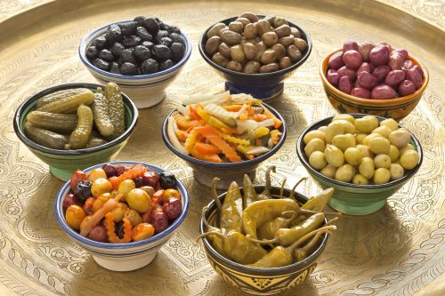 A decorated tray holds an assortment of small bowls, each holding a different preserved food such as olives, chili peppers, cornichons and marinated vegetables. 