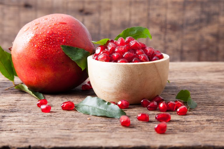 A small bowl full of juicy pomegranate seeds. A few seeds are scattered around the base of the bowl and a whole unopened pomegranate is in the background.