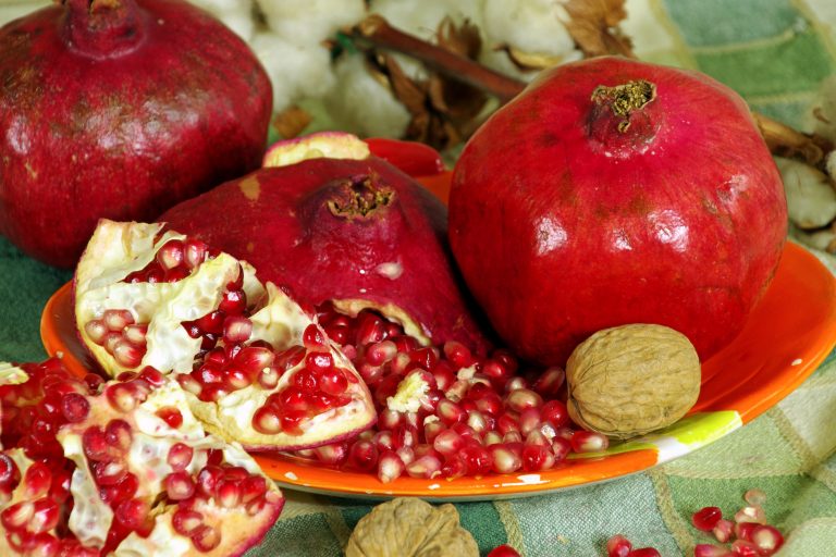 Image showing several wedges of pomegranate; the bright red seeds are exposed but still trapped in membrane. Several whole pomegranates are in the background. A single whole walnut rests beside a small pile of pomegranate seeds.