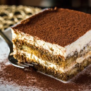 A square-shaped serving of tiramisu, an Italian dessert made by layering coffee-dipped ladyfingers with a mascarpone cream cheese mixture. The top of the dessert is heavily dusted with cocoa powder.