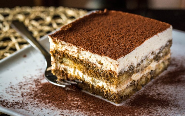 A square-shaped serving of tiramisu, an Italian dessert made by layering coffee-dipped ladyfingers with a mascarpone cream cheese mixture. The top of the dessert is heavily dusted with cocoa powder.