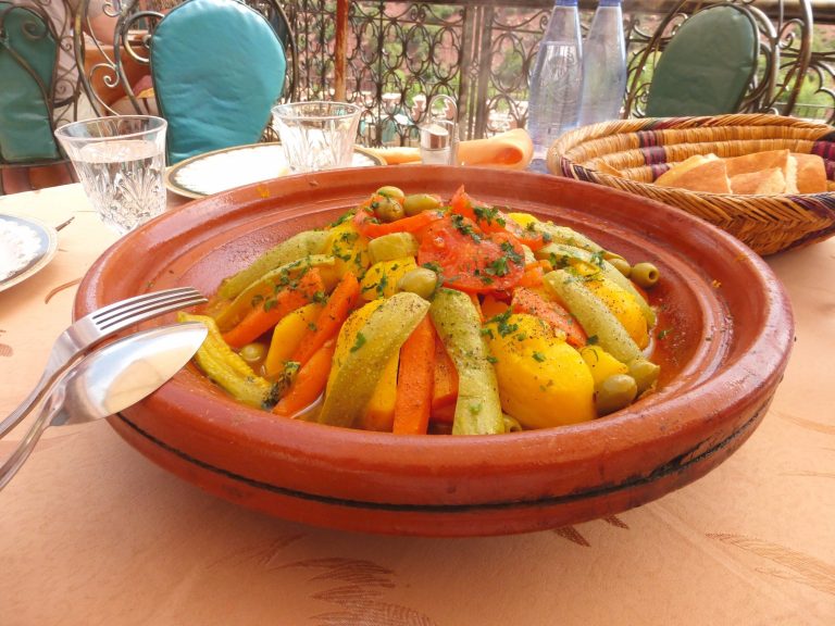 A close-up image of a Moroccan tagine. An assortment of vegetables including carrots, zucchini and potatoes are arranged in pyramid fashion. The dish is garnished with green olives and tomatoes.
