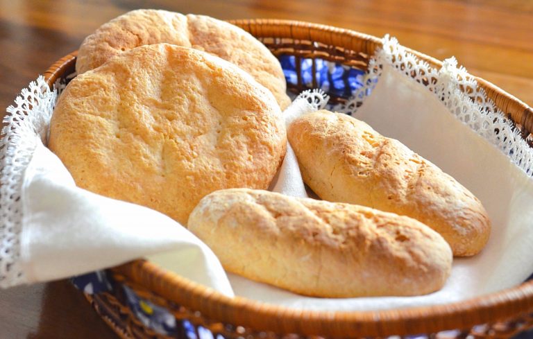 Photo of gluten-free Moroccan bread. Two gluten-free round loaves and two gluten-free mini-baquettes sit on a white linen napkin in a bread basket on a wooden table.