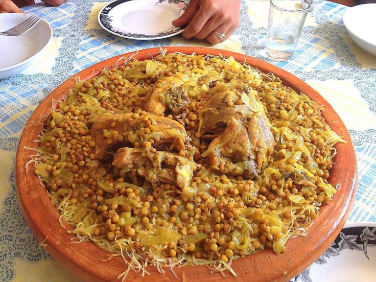 A large, deep ceramic dish holding a Moroccan dish of chicken, lentils and onions served on a bed of shredded, stringy pan-fried crepes called rezzat el qadi.