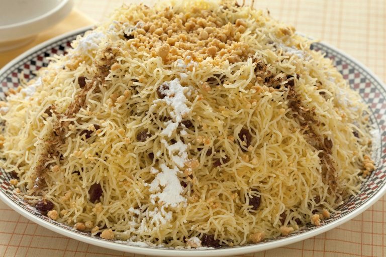 Steamed, broken strands of vermicelli are mounded high on a platter and decorated ground fried almonds, cinnamon and powdered sugar. Raisins can be seen throughout the vermicelli.