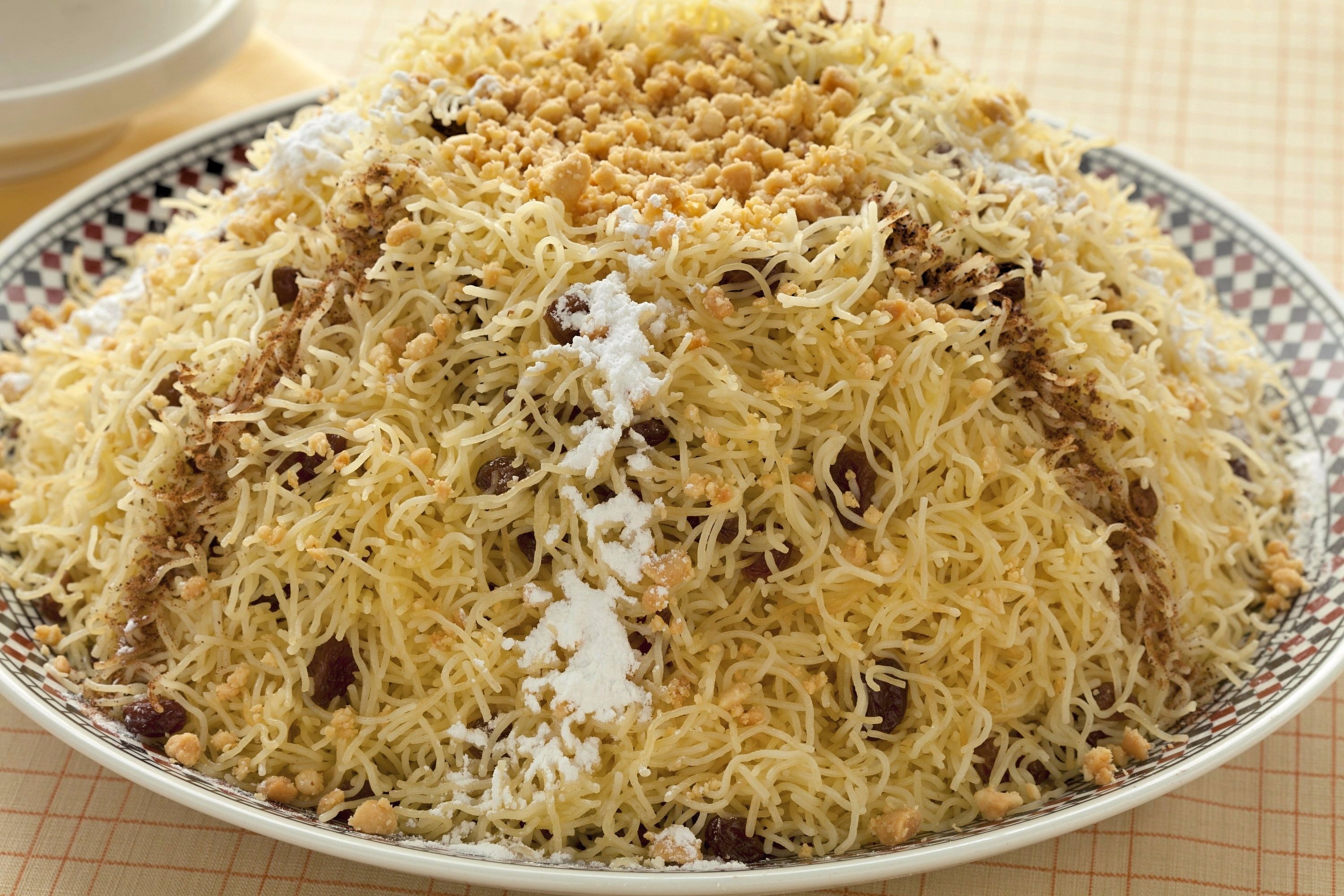 Steamed, broken strands of vermicelli are mounded high on a platter and decorated ground fried almonds, cinnamon and powdered sugar. Raisins can be seen throughout the vermicelli.