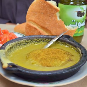 Moroccan split pea soup in a black shallow metal dish. The soup is garnished with ground cumin and olive oil. A spoon is in the soup and a partial loaf of Moroccan bread and bottle of olive oil are behind the bowl.