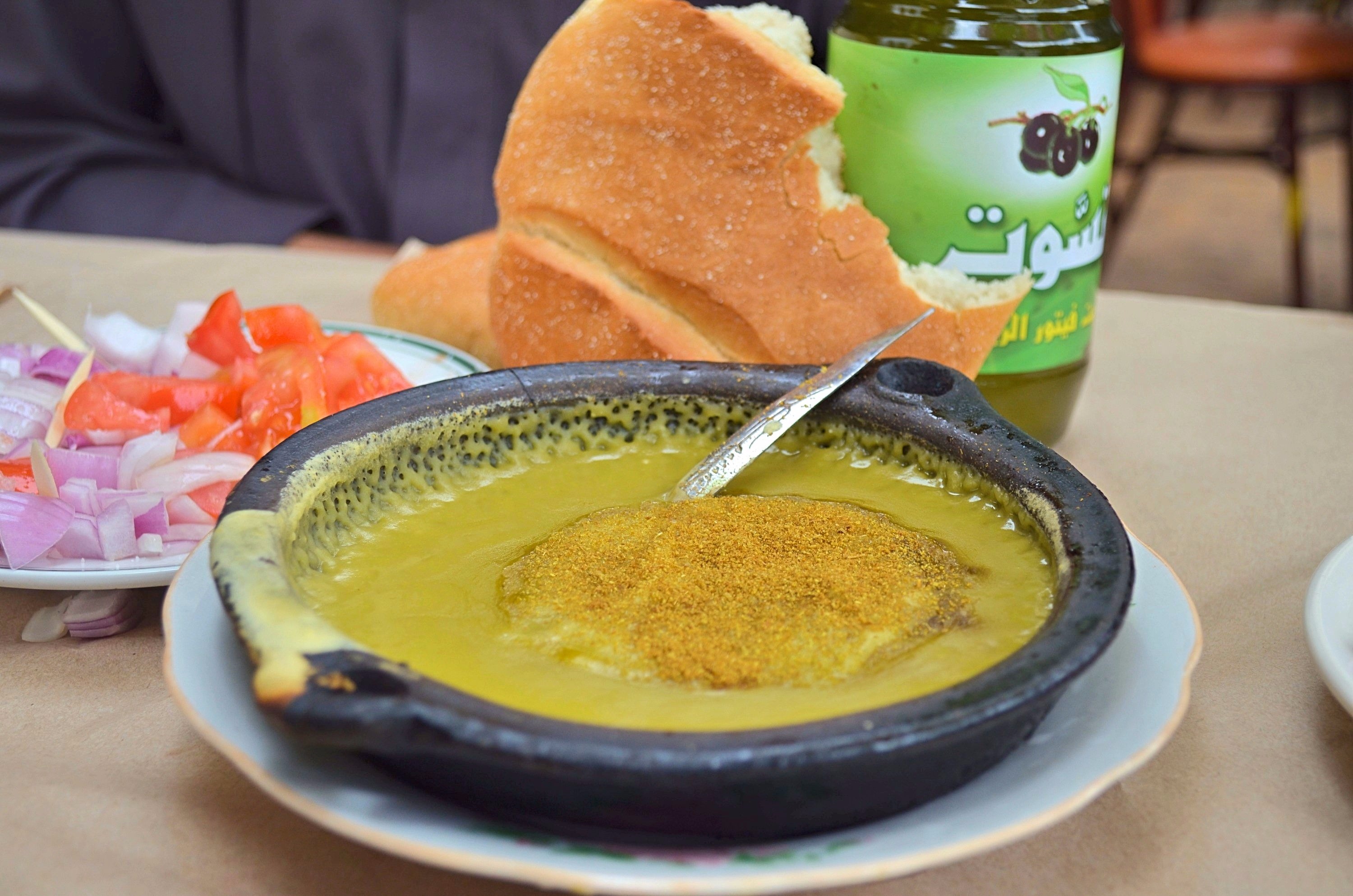Moroccan split pea soup in a black shallow metal dish. The soup is garnished with ground cumin and olive oil. A spoon is in the soup and a partial loaf of Moroccan bread and bottle of olive oil are behind the bowl.