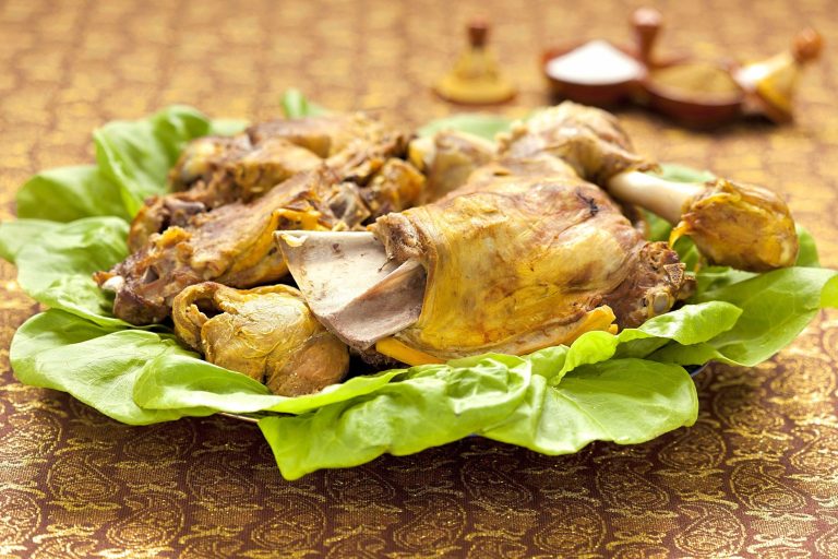 A lettuce-line platter holds large pieces of steamed lamb. Small tagine-shaped condiment dishes holding salt and cumin are in the background.