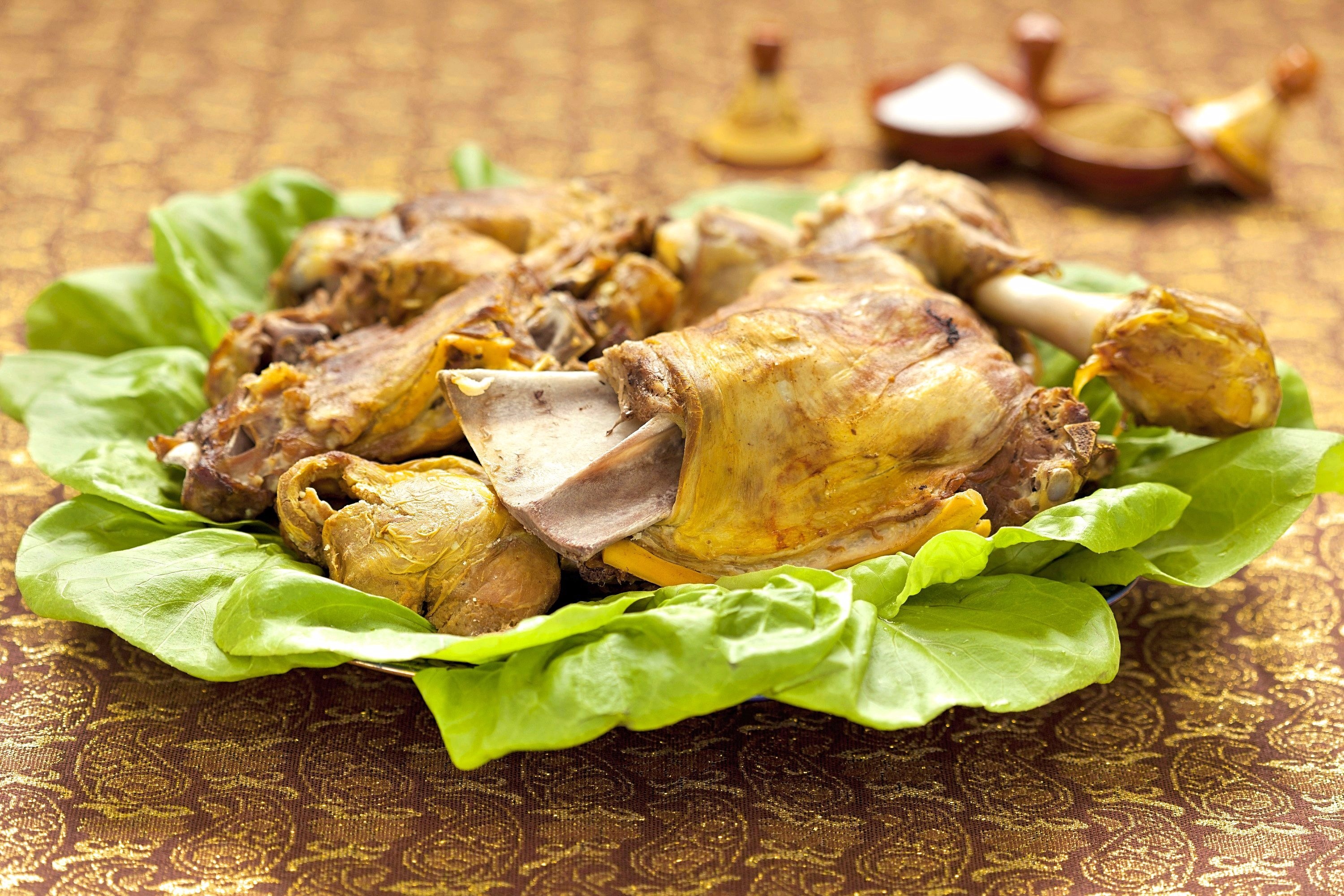 A lettuce-line platter holds large pieces of steamed lamb. Small tagine-shaped condiment dishes holding salt and cumin are in the background.