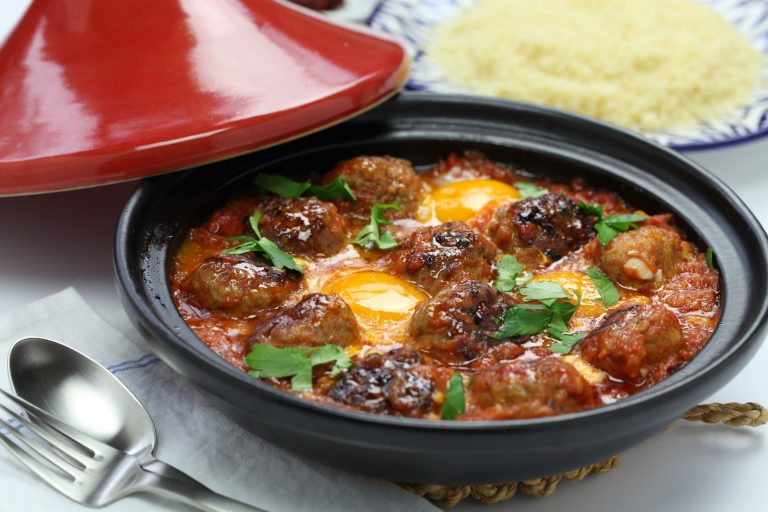 Small Moroccan meatballs and poached eggs in tomato sauce sauce are ready to be served from a tagine. The dish is garnished with fresh parsley and part of the red conical lid can be seen resting against the tagine base.