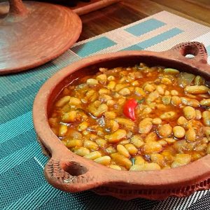 A shallow clay dish with handles holds Moroccan loubia, a dish of white beans stewed in zesty tomato sauce. A small red chili pepper is centered in the dish.