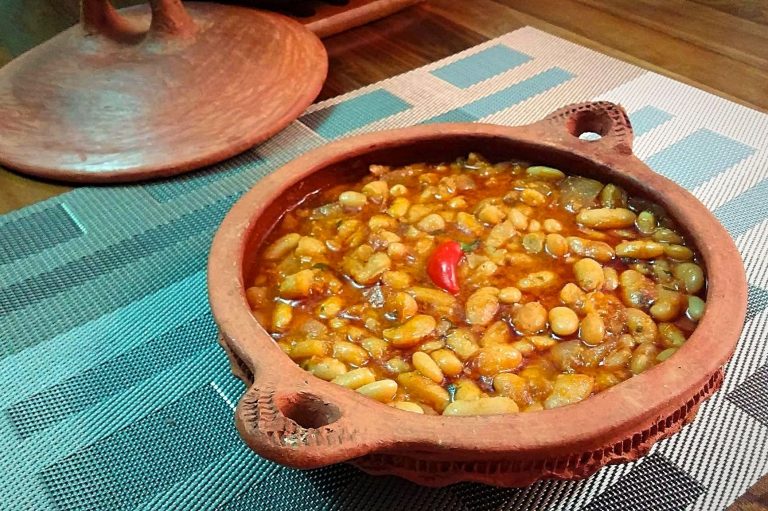 A shallow clay dish with handles holds Moroccan loubia, a dish of white beans stewed in zesty tomato sauce. A small red chili pepper is centered in the dish.
