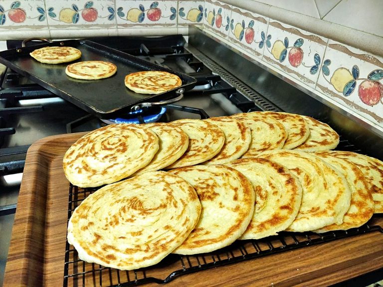 Freshly-made round Moroccan pancakes called meloui are on a cooling rack set on a tray. Three of the pancakes can be seen cooking on a griddle in the background.