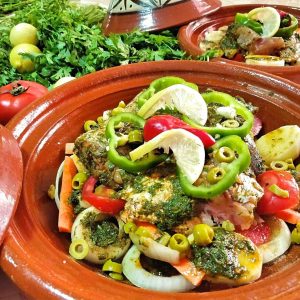 A tagine base is filled with carefully arranged fish and vegetables. In the background are another prepped fish tagine as well as fresh herbs and lemon.