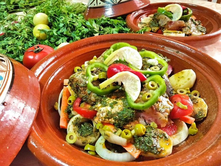 A tagine base is filled with carefully arranged fish and vegetables. In the background are another prepped fish tagine as well as fresh herbs and lemon.