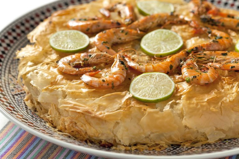 Close-up photo of Moroccan Seafood Bastilla, a savory seafood pie wrapped in crispy paper-thin pastry. The pie is garnished with cooked shrimp and thin slices of lemon.