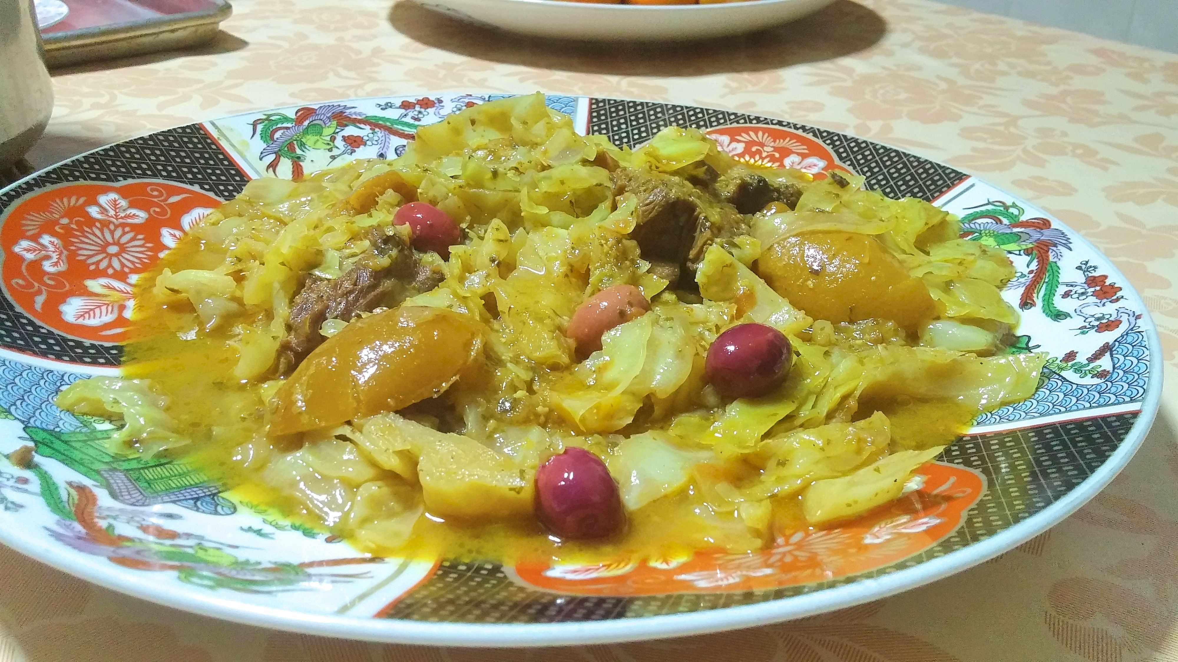 Moroccan stewed cabbage is served on a decorated plate. Pieces of meat can be seen in the cabbage, and the dish is decorated with wedges of preserved lemon and olives.