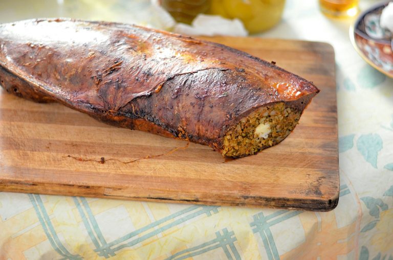 A baked stuffed veal spleen sits on a wooden cutting board. A slice has been cut from the end facing the camera, revealing a filling of kefta, rice and egg.