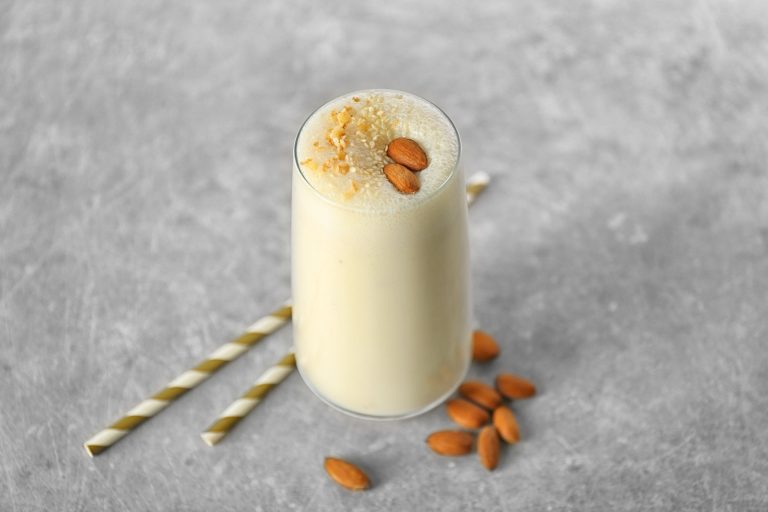 A glass of almond milkshake sits on a table next to two straws and several scattered almonds.