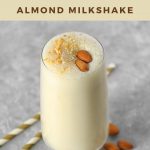 A glass of almond milkshake sits next to two straws and a few scattered almonds.